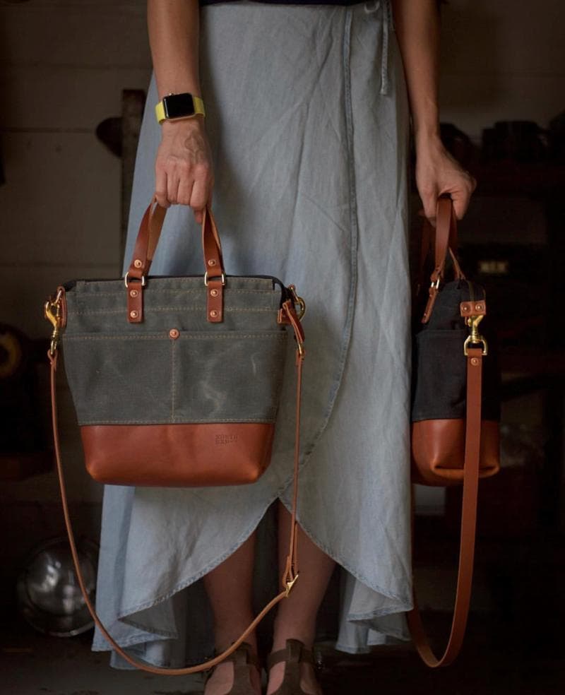 Person holding two leather and fabric bags, one in each hand. The bags have brown leather details and adjustable straps. The person is wearing a light blue skirt and a smartwatch.
