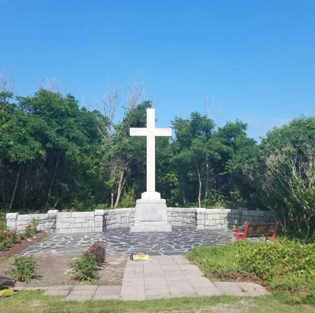 A large white cross stands on a stone platform surrounded by shrubs and trees. A red bench is nearby, under a clear blue sky.