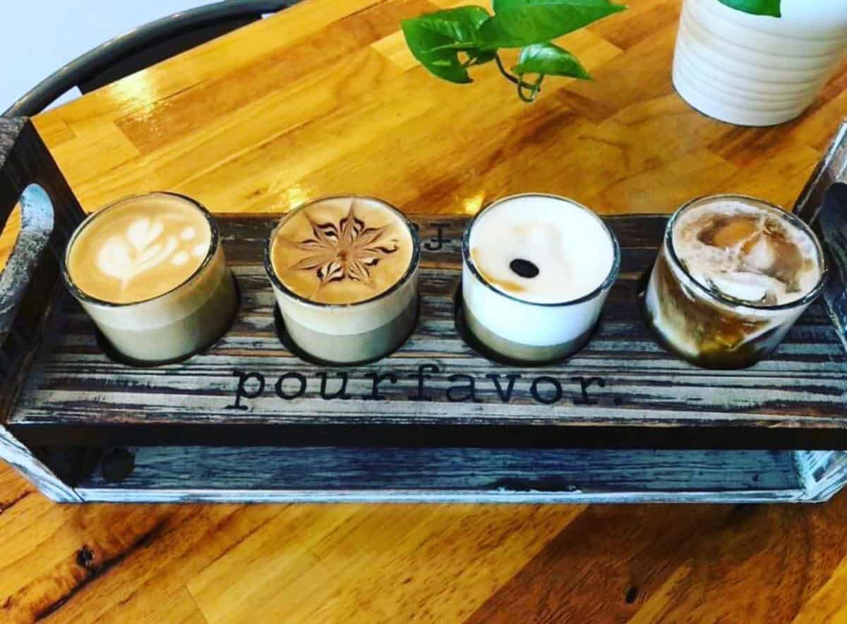 A wooden tray with four varieties of coffee, each in a glass cup with different latte art, on a wooden table with a small plant in the background.
