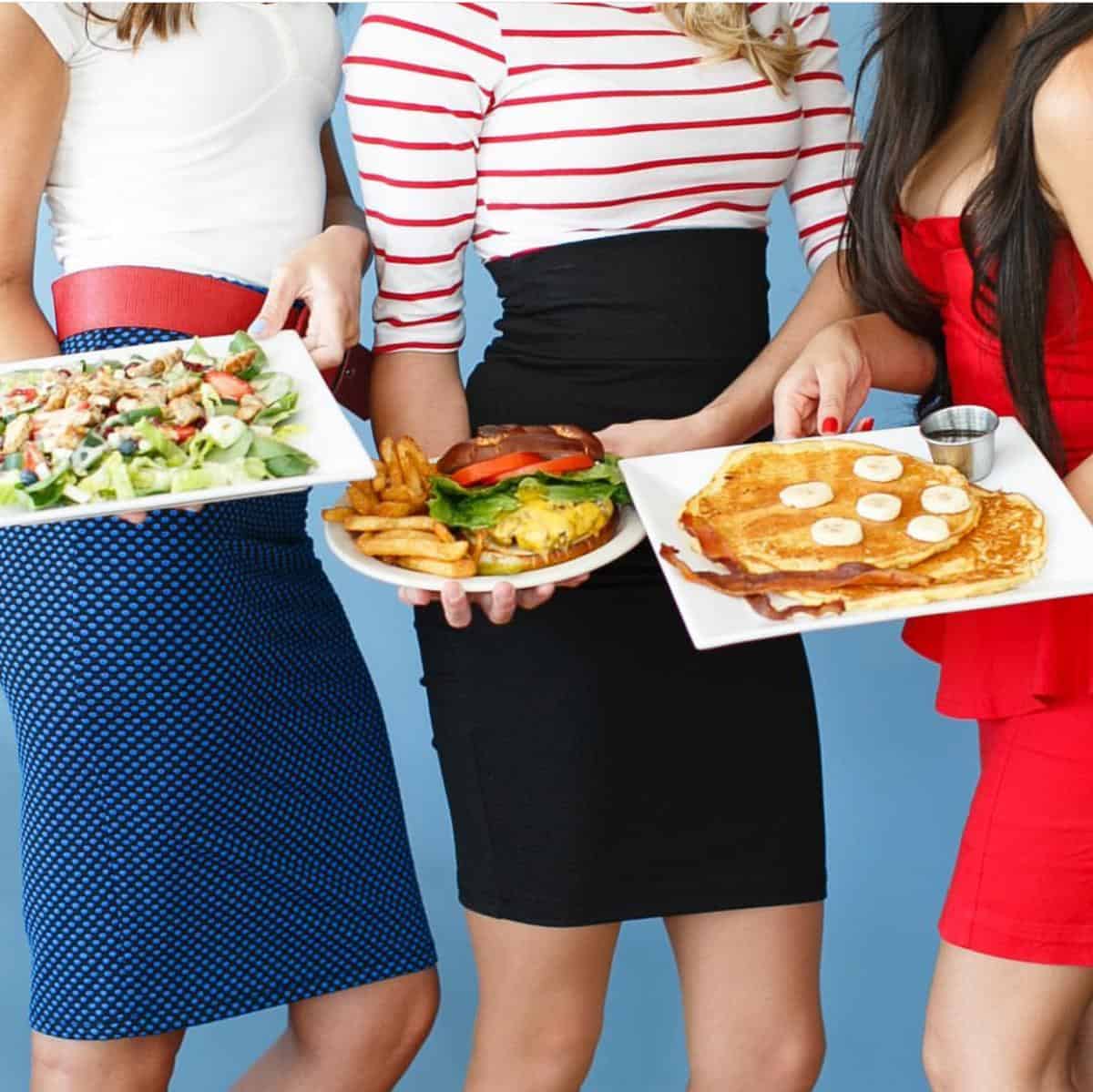 Three people holding plates with a salad, burger with fries, and pancakes with banana slices against a blue background.