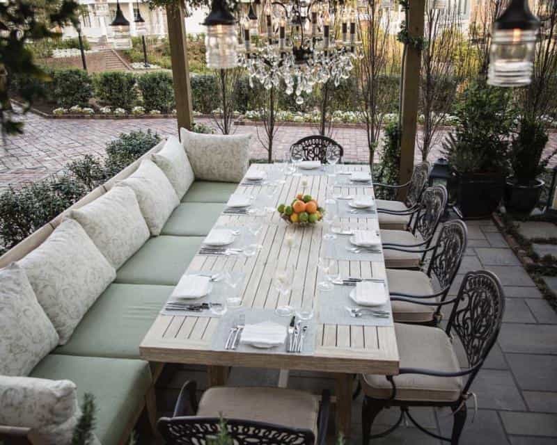 Outdoor dining area with a long table set for eight, featuring white plates and cutlery. A fruit bowl is at the center, surrounded by cushion seating and ornate chairs under hanging lights.