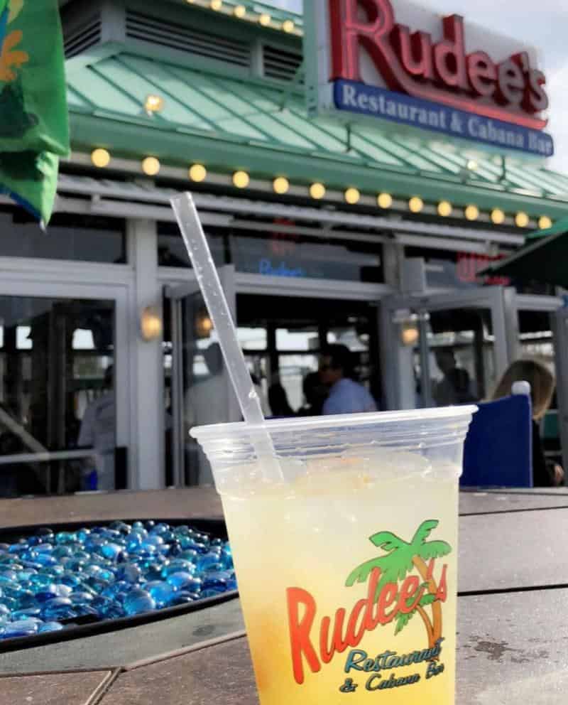Plastic cup with a yellow beverage and straw on a table, in front of Rudee's Restaurant & Cabana Bar entrance. Blue stones visible on table surface.