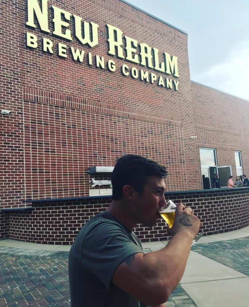 A person drinks from a glass outside New Realm Brewing Company, a brick building with a sign on the facade.