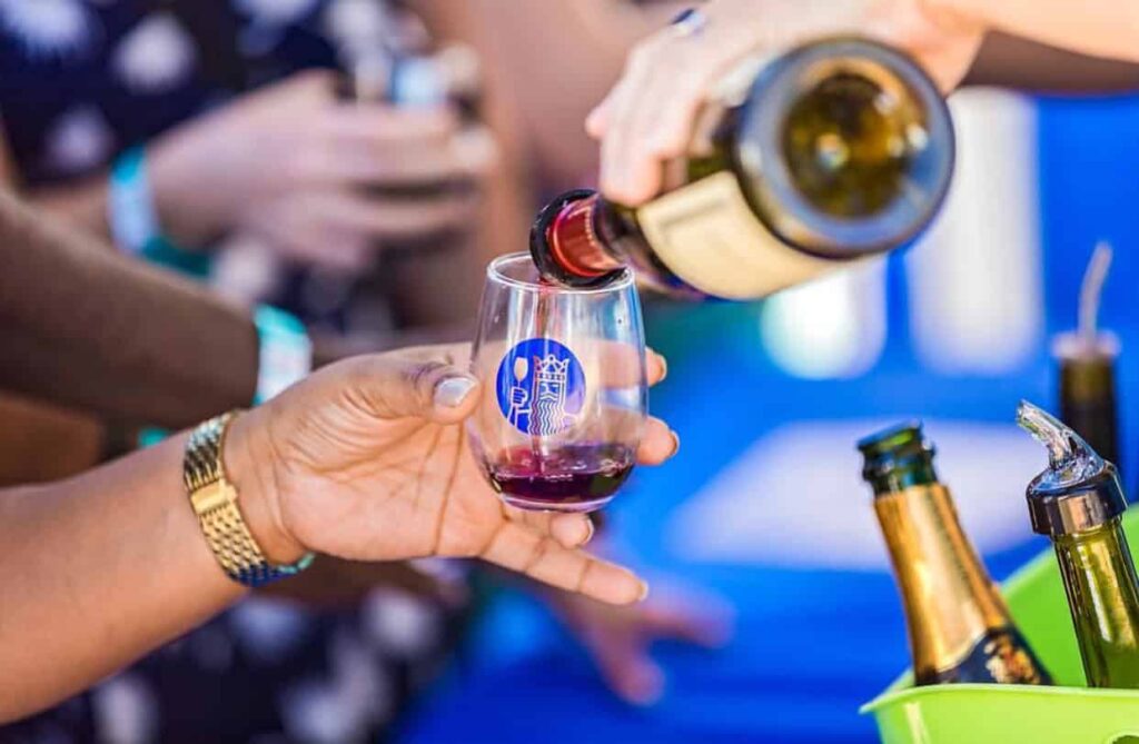 A person pours red wine into a glass held by another hand. The scene includes bottles and is set outdoors with people in the background.