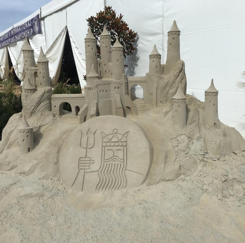 Elaborate sandcastle sculpture with multiple towers and an engraved image of a bearded figure holding a trident, set against a white tent background.