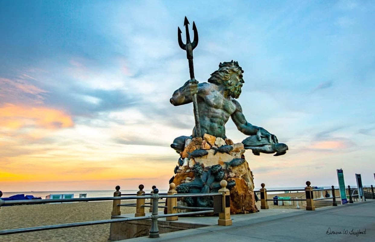 Large statue of a muscular figure wielding a trident, resembling Neptune, situated on a boardwalk with a beach and sunrise in the background.