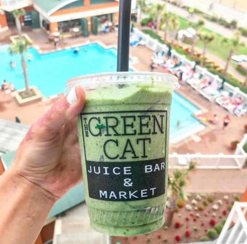 A hand holds a green smoothie in a cup labeled "The Green Cat Juice Bar & Market" against a backdrop of a pool area with palm trees and lounge chairs.