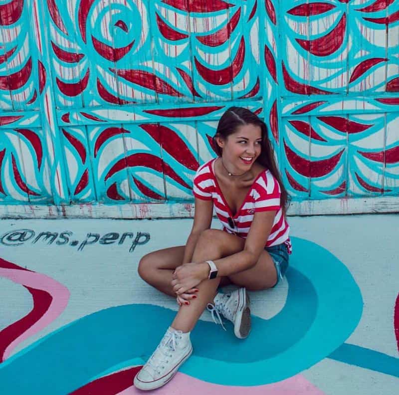 Woman in a red and white striped shirt sits on a colorful, abstract painted surface, smiling. Blue and red swirls decorate the background.