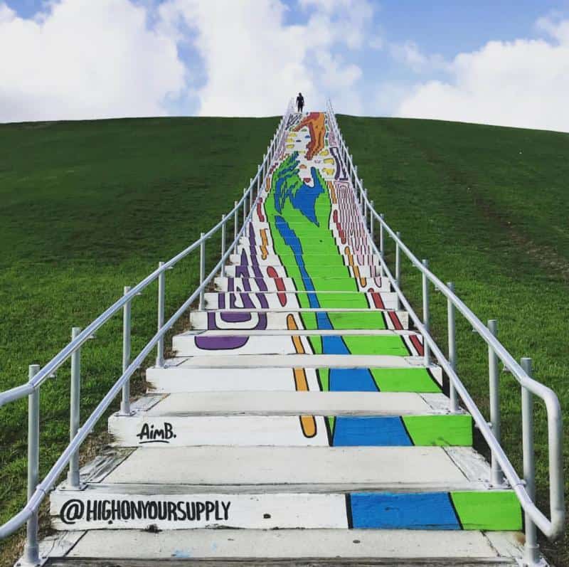 Colorful mural of a woman painted on outdoor staircase with handrails, set against a grassy hill and blue sky.
