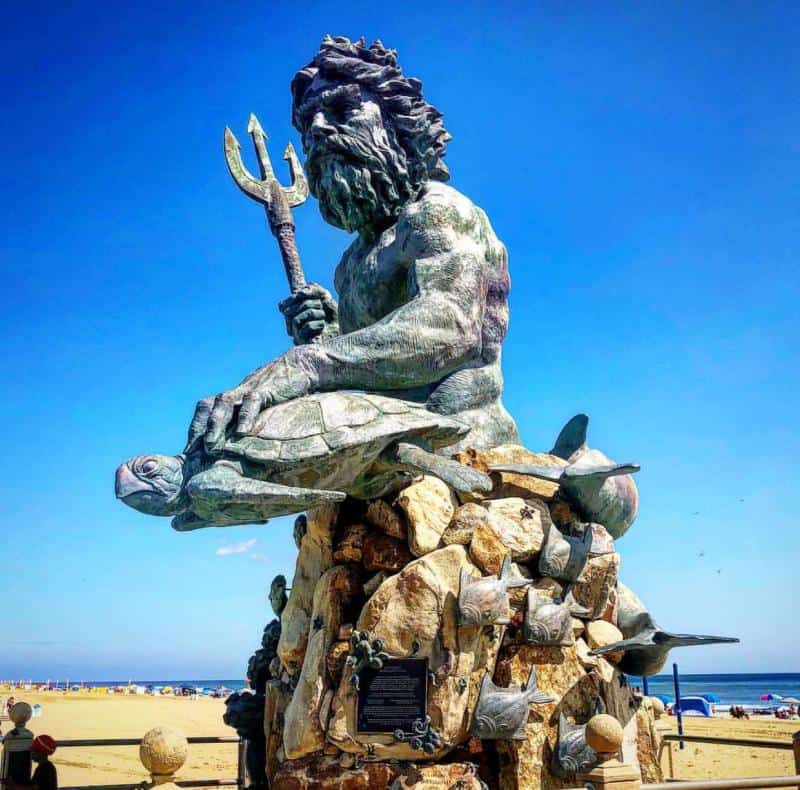 A large bronze statue of Poseidon holding a trident, surrounded by marine creatures, stands on a beach against a clear blue sky.