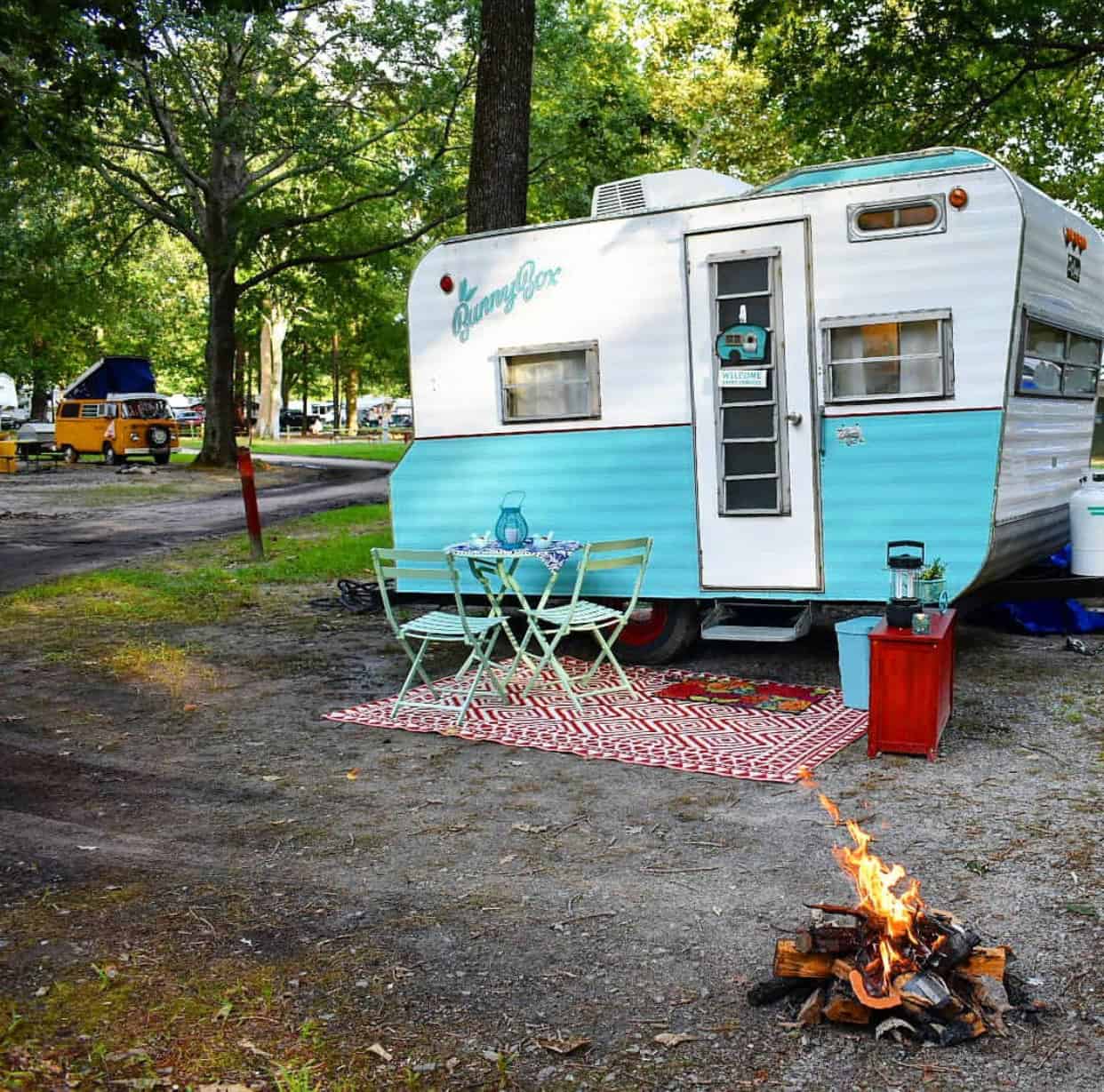 Vintage blue and white camper with a small outdoor setup, including a table and chairs on a patterned rug. A campfire burns nearby, surrounded by tall trees.