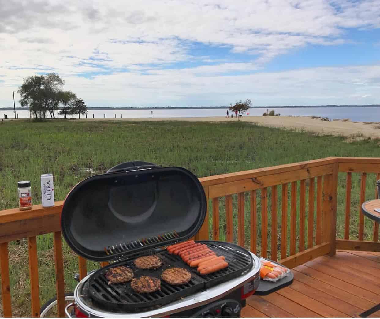 Grill on a wooden deck holding burgers and hot dogs. Scenic view of a grassy field and sandy beach with a blue sky and clouds in the background.