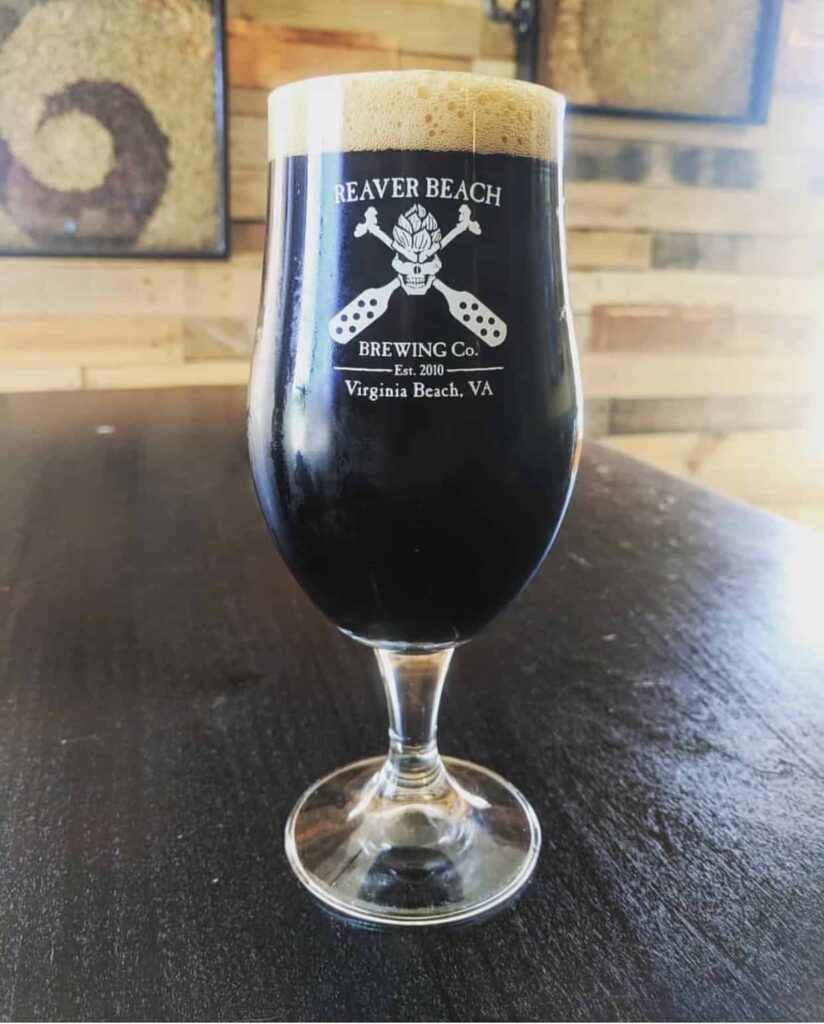 A glass of dark beer from Reaver Beach Brewing Co., Virginia Beach, VA, is on a dark wooden table with a blurred background.