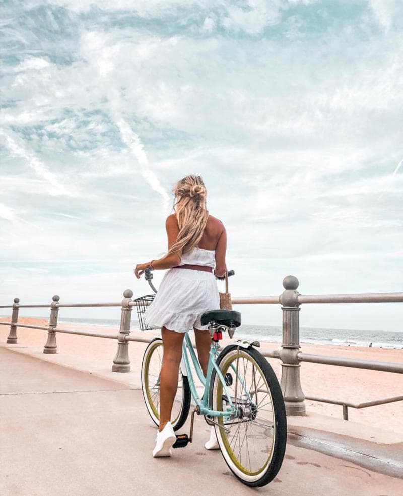 Person with long hair, in a white dress, stands beside a bicycle on a seaside walkway, facing the ocean under a partly cloudy sky.