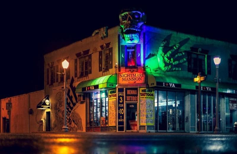 A brightly lit storefront named "Nightmare Mansion" with a large skull and claws, offering tattoos and henna, located on a street corner at night.
