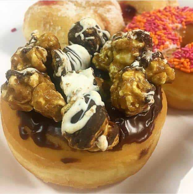 A donut topped with chocolate icing and caramel popcorn. Other donuts with sprinkles and filling are blurred in the background.