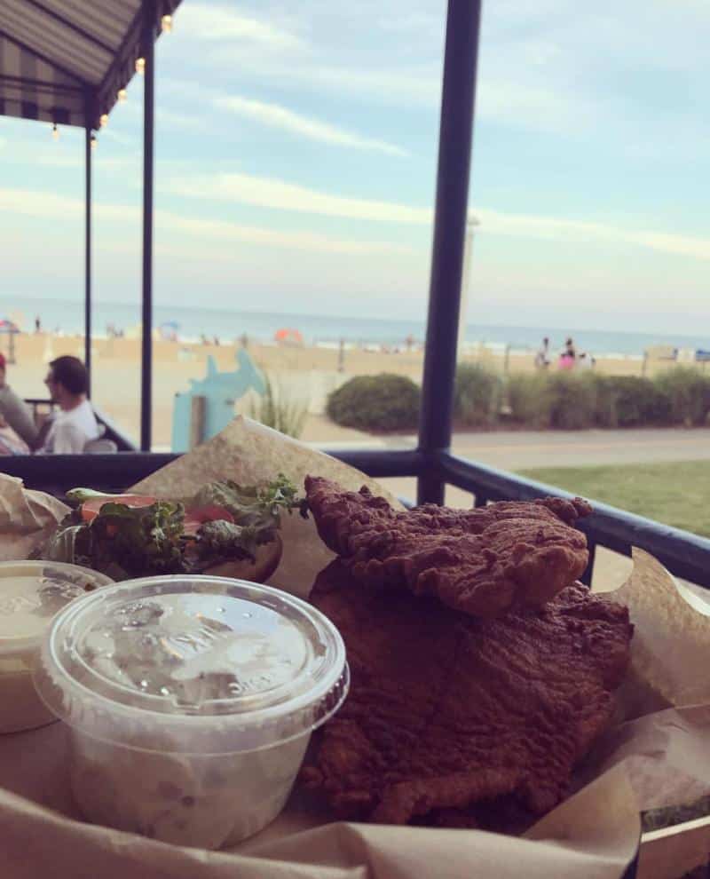 Fried fish with sauce and salad in a basket, overlooking a beach scene with sand, sea, and sky in the background.