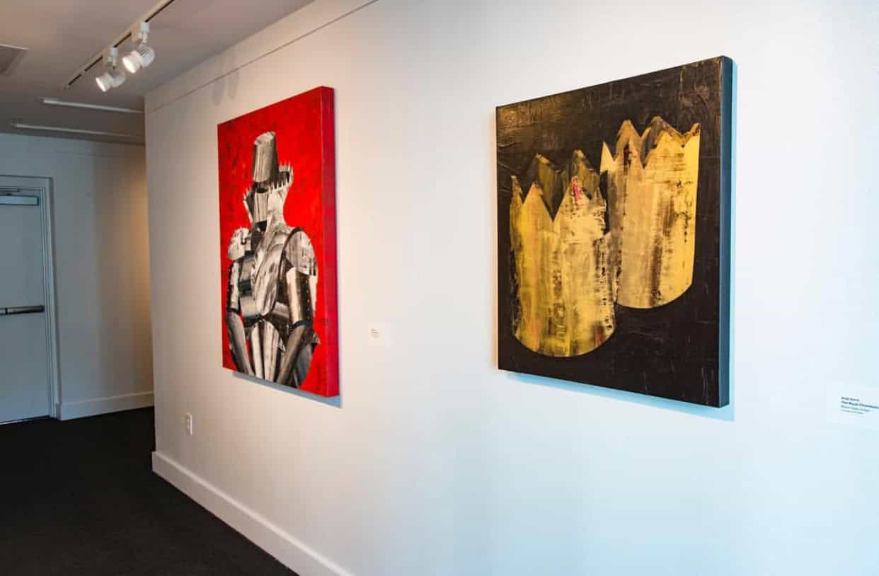 Two paintings hang on a white gallery wall. The left painting features a figure on a red background, while the right painting displays abstract crown-like shapes in gold and black.