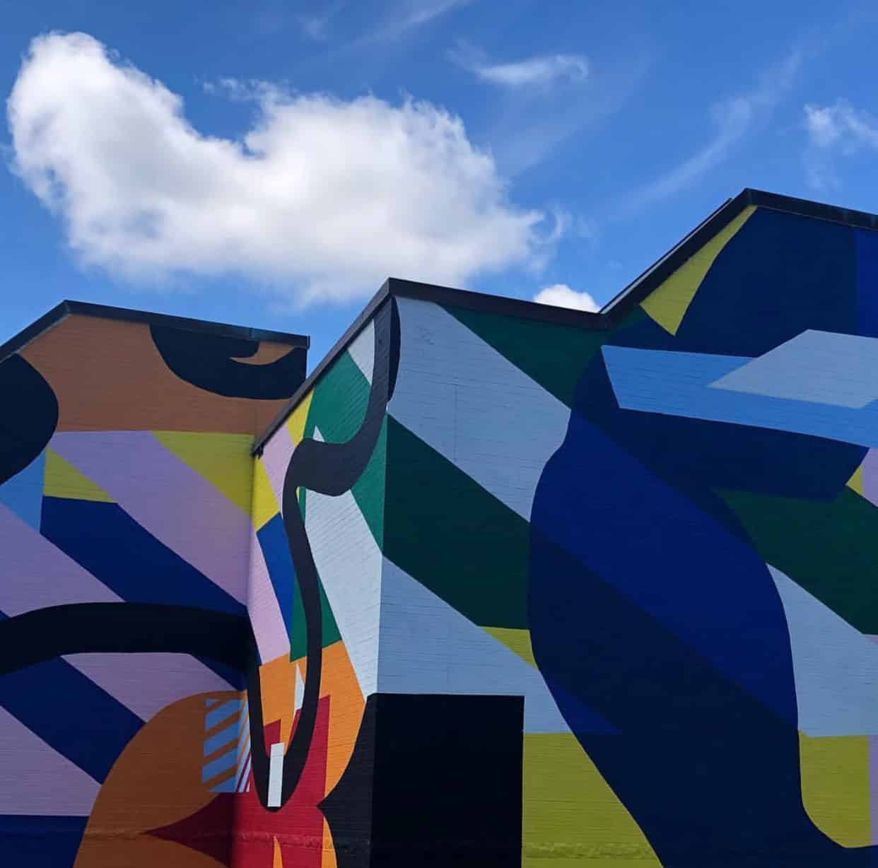 Colorful abstract mural with geometric shapes and bold patterns on a building. Bright blue sky with a few clouds in the background.