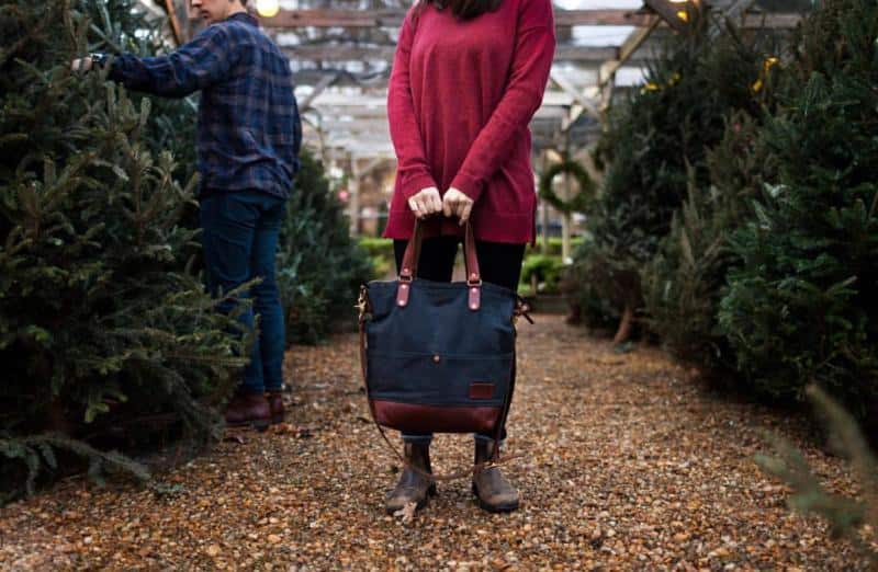 Person holding a black and brown bag stands on a gravel path between Christmas trees. Another person in a plaid shirt is examining trees in the background.