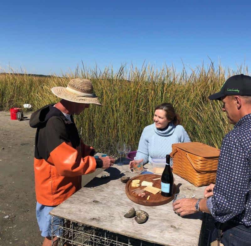 Three people stand by a makeshift table with cheese, wine, and shells in a grassy outdoor setting under a clear blue sky.