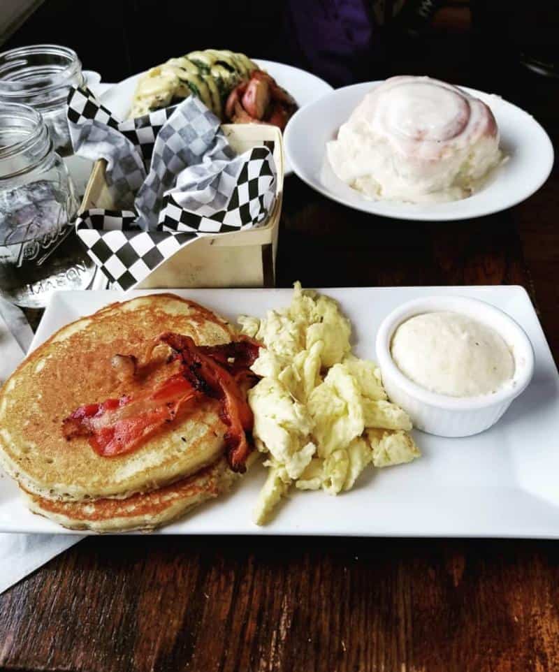 A breakfast plate with pancakes, bacon, scrambled eggs, and a small dish of butter. A cinnamon roll and other dishes are in the background.