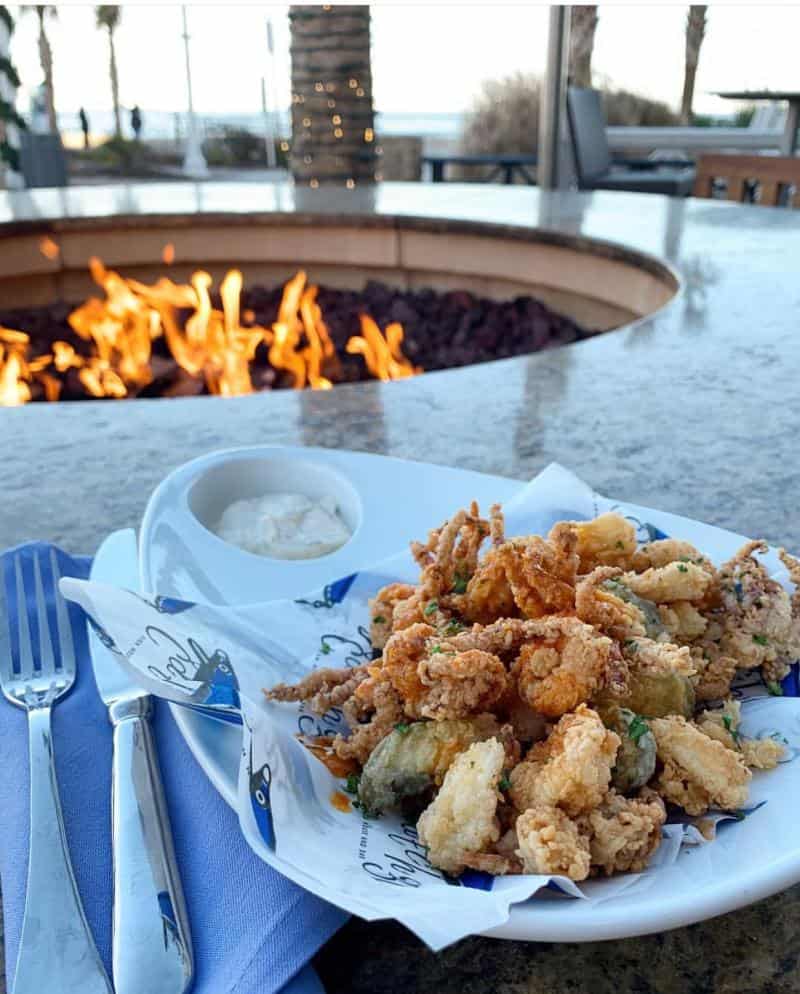 A plate of fried calamari with dipping sauce on a table near a fire pit, with a fork and napkin beside it.