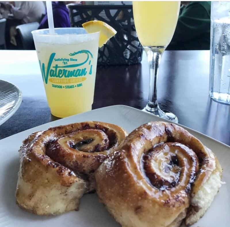 Two cinnamon rolls on a white plate next to a drink in a branded cup and a tall glass with a yellow beverage.