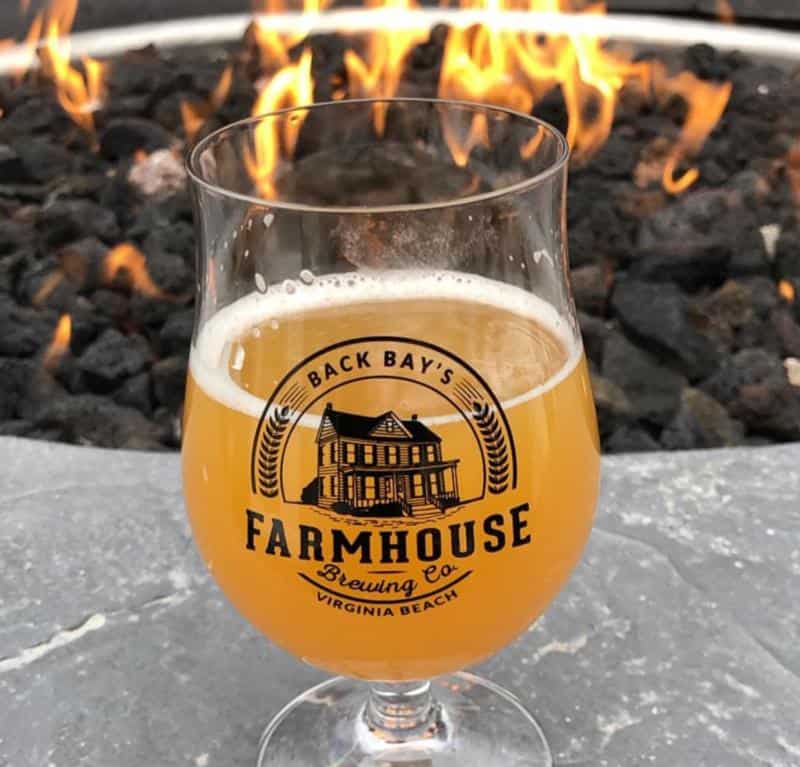 A glass of beer labeled "Back Bay's Farmhouse Brewing Co." is placed on a stone surface in front of a fire pit with flames.