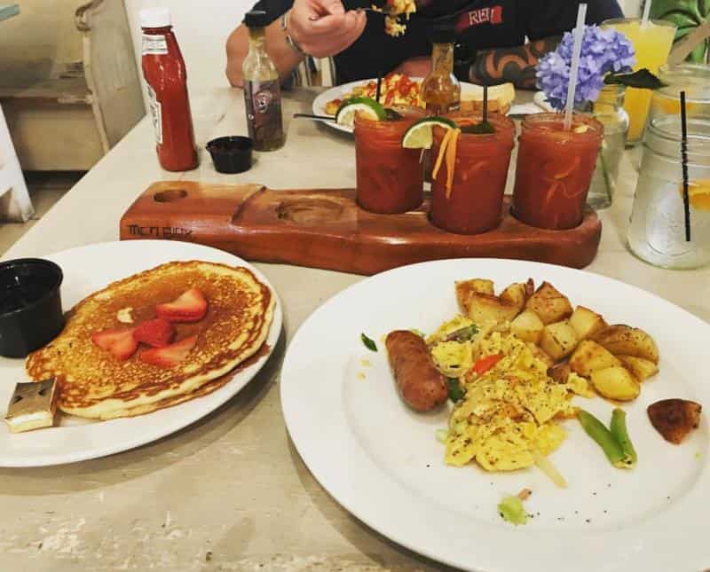 A breakfast table with pancakes topped with strawberries and butter, scrambled eggs, sausage, potatoes, and Bloody Mary cocktails.