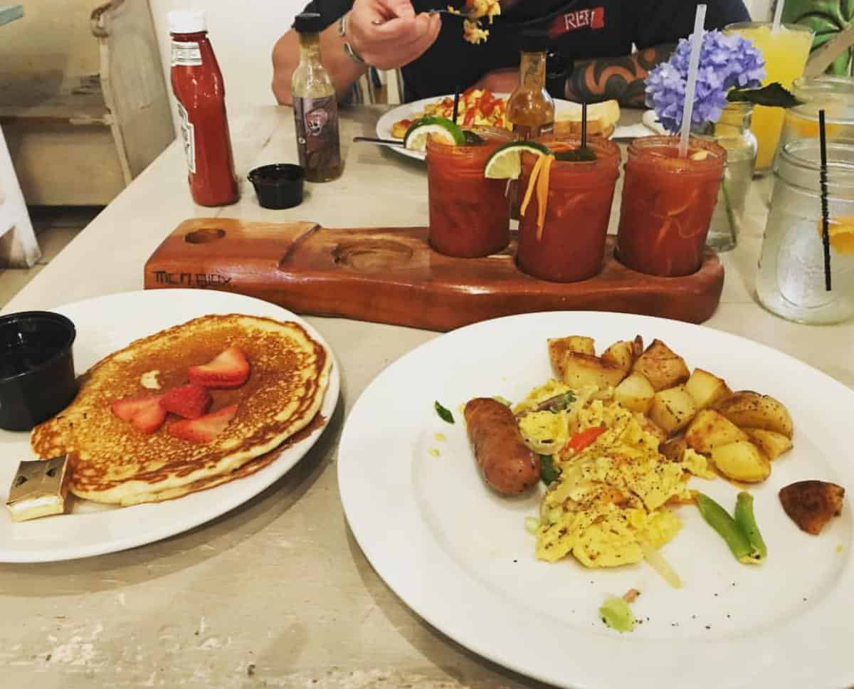 Pancakes with strawberries and butter, scrambled eggs with sausage and potatoes, three Bloody Mary drinks, ketchup and hot sauce bottles on a table.