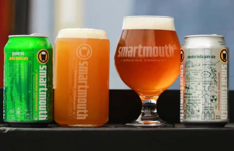 Four Smartmouth beer products, including two cans and two filled glasses, displayed on a surface.