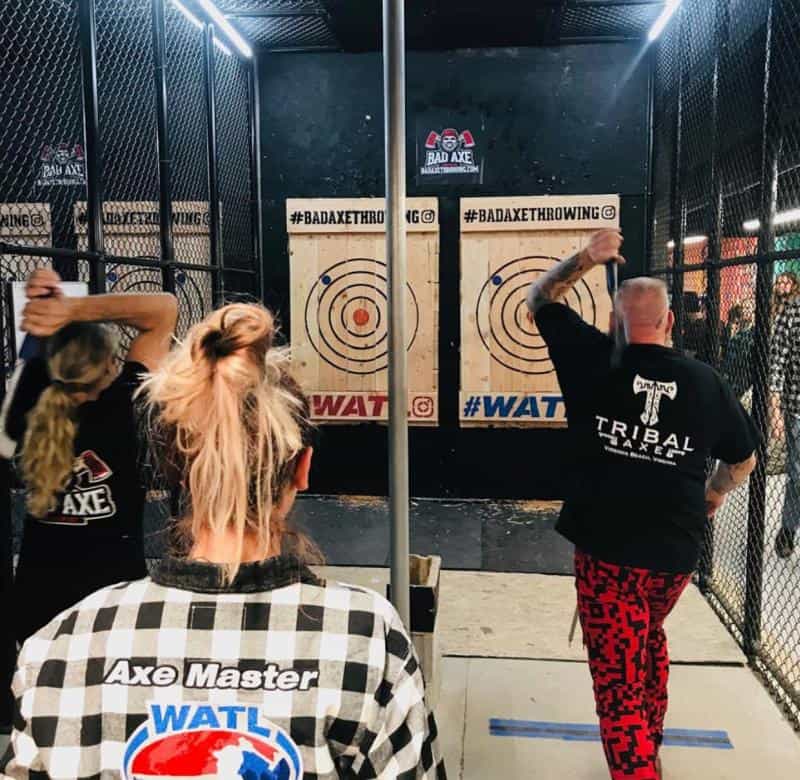 Two people in an axe-throwing venue aim at wooden targets. One wears a "Tribal" shirt with red pants, and another has a checkered shirt with "Axe Master" on the back.