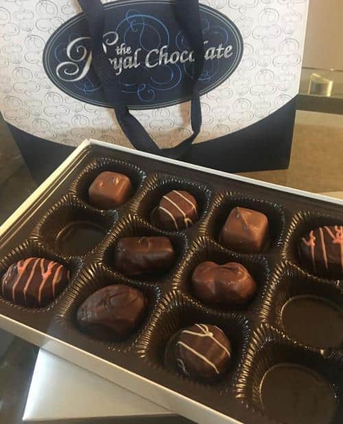 A box of assorted chocolates with decorative toppings, open in front of a branded bag from The Royal Chocolate.