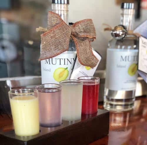 Four glasses of different colored liquids are in a row. Behind them are bottles labeled "Mutiny Island" adorned with burlap bows.