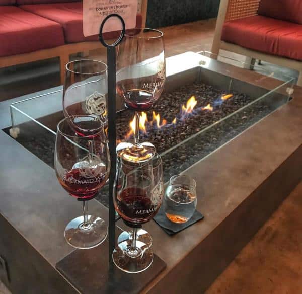 Four wine glasses on a metal holder and a glass of whiskey are placed on a table next to a glass-enclosed fire pit with visible flames.