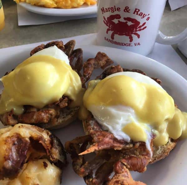 Eggs Benedict with crab, topped with hollandaise sauce, served alongside a stuffed potato and a mug with "Margie & Ray's Sandbridge, VA" logo.