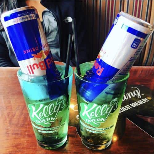 Two glasses with Kelly's Tavern branding each contain a partially submerged Red Bull can and a straw.