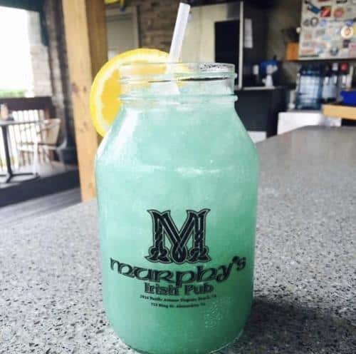 Jar of blue-green lemonade with ice and a lemon slice, served in a glass with a straw on a bar counter.