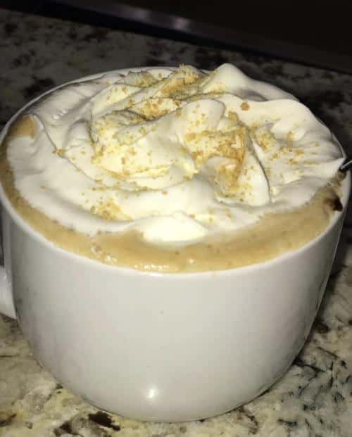 A white mug filled with a frothy hot drink topped with whipped cream and sprinkled with a light dusting of cinnamon or spice.
