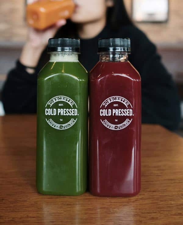 Two bottles of cold-pressed juice, one green and one red, on a wooden table. A person in the background drinks from an orange bottle.