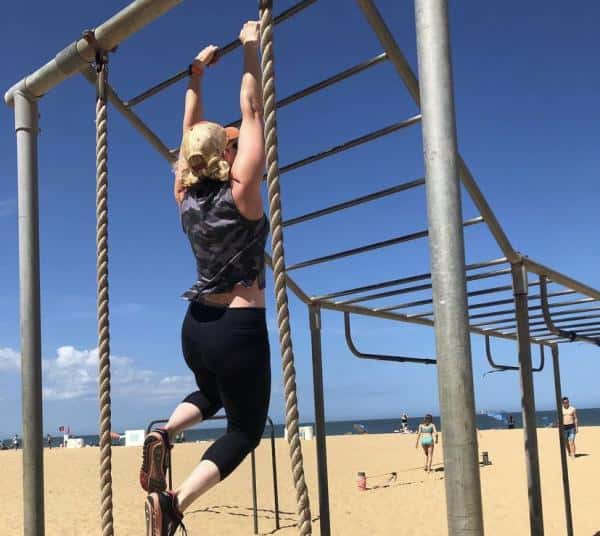 Person climbing a rope on a metal structure at a beach. Others are visible in the background.
