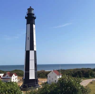 A black and white lighthouse stands near the coast, surrounded by greenery and buildings, with the ocean visible in the background under a clear blue sky.