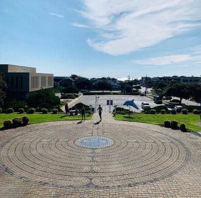 Wide view of a brick circular plaza with a pattern, surrounded by a grassy area. Three people walk in the distance under a partly cloudy sky. Buildings and a parking lot are visible.