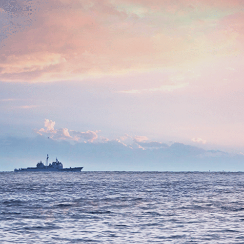 A naval ship sails on a vast ocean under a pastel sky during sunset.