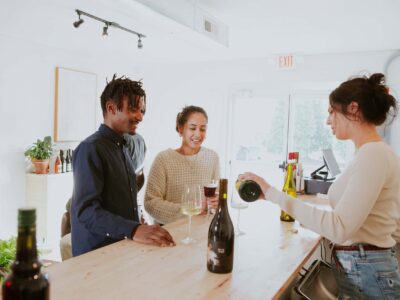 Three people at a wine bar: a server pours wine for two smiling patrons, one with a clear drink and one with a dark drink. Bottles and glasses are on the wooden counter.
