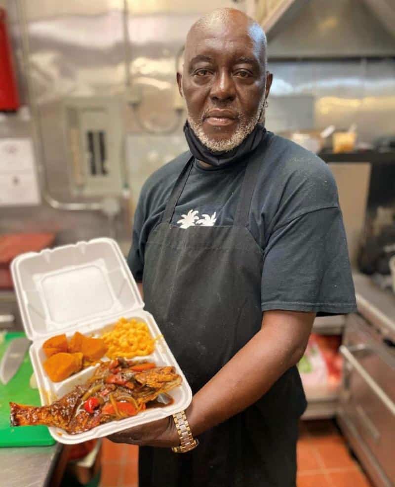 A man in a kitchen holds a takeout container with fish, plantains, and macaroni.