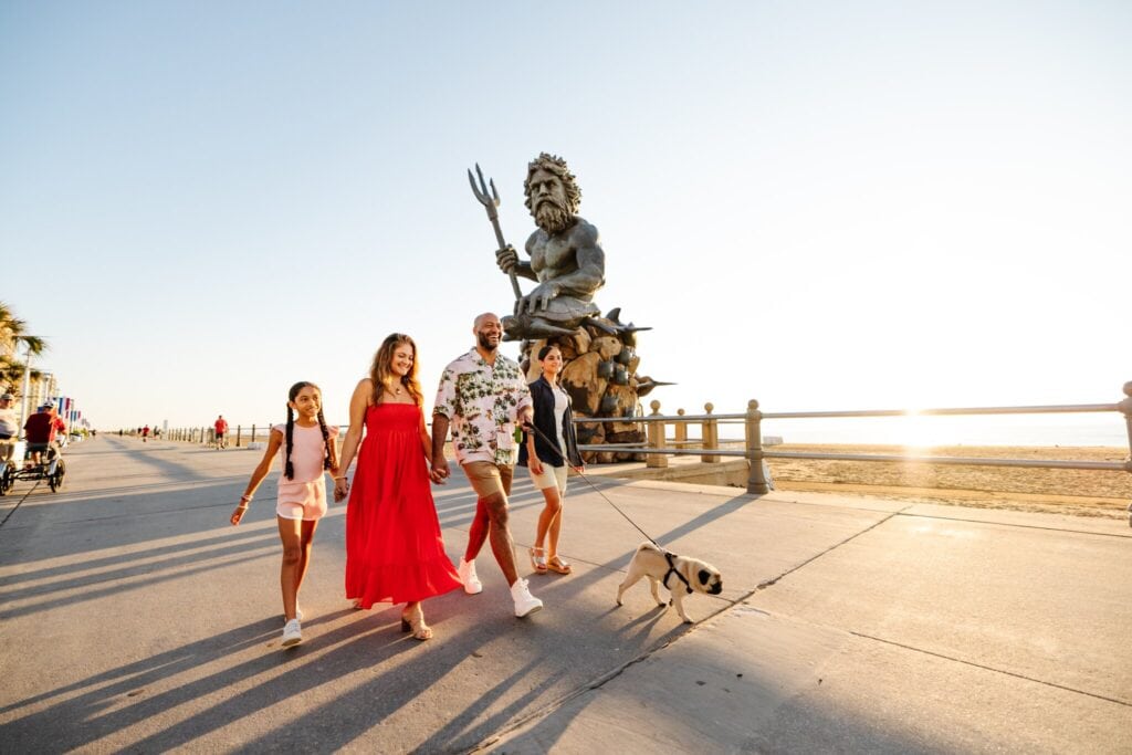 A family strolls along a beachfront promenade with a dog, passing by the King Neptune statue at sunset.