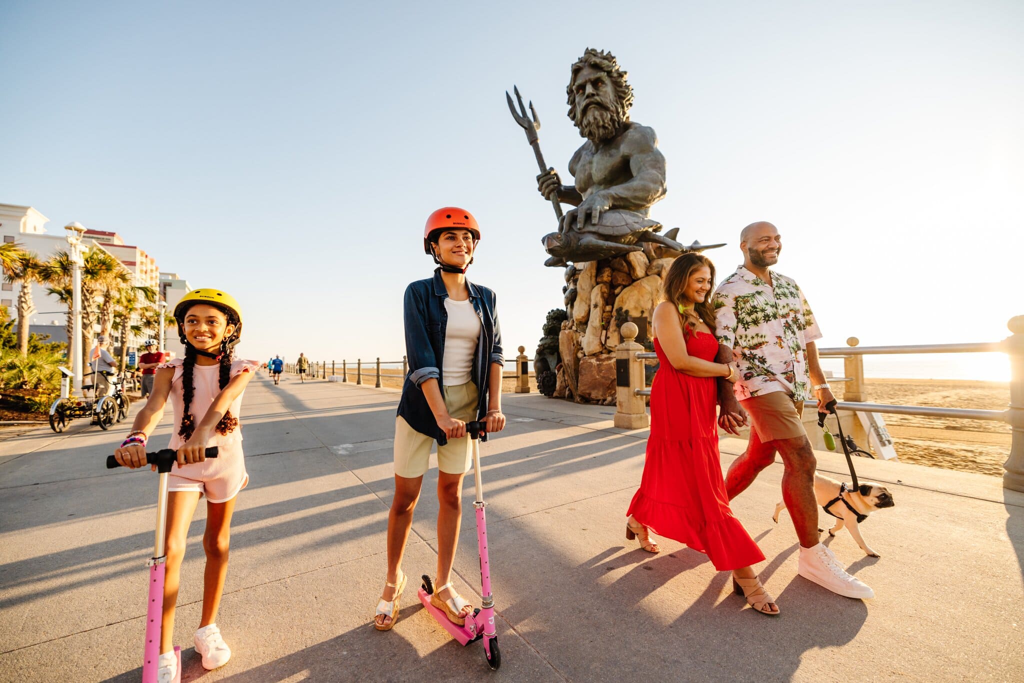 A girl and a young woman ride scooters along a beachside walkway, while a couple walks hand in hand with a dog, near a large statue.