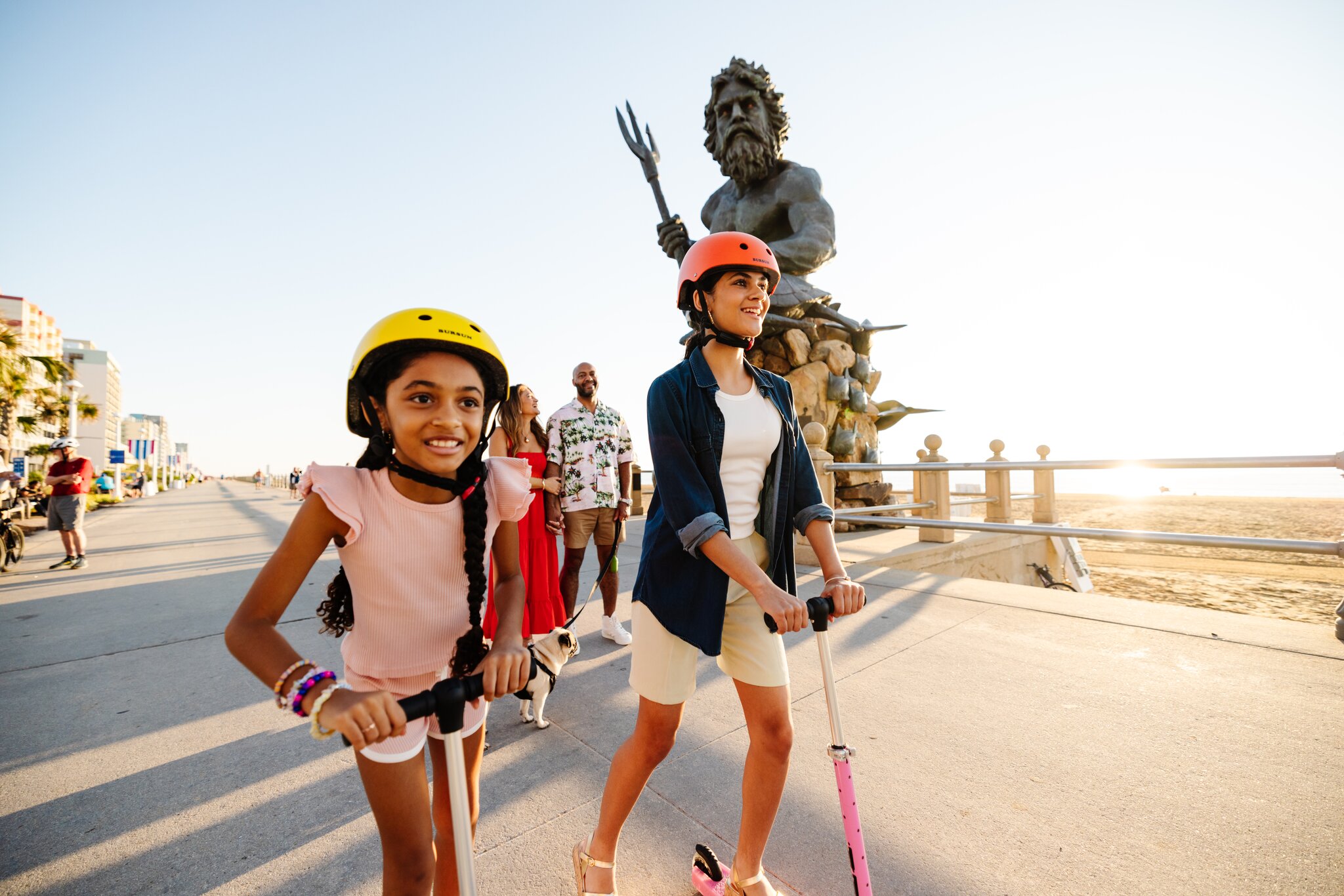 Two girls riding scooters on a boardwalk near a beach, with a large statue of Neptune in the background during sunset.